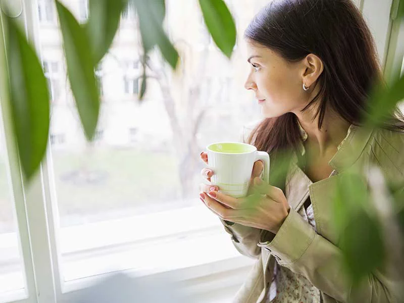 A woman gazes out the window while holding a cup of tea, reflecting a moment of calm and contemplation.