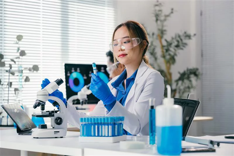 Young scientist wearing lab coat and gloves analyzing test tube and holding microscope while sitting at desk in modern laboratory