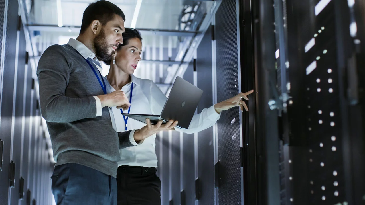 Two people focused on a laptop in a server room, surrounded by rows of servers and technical equipment.
