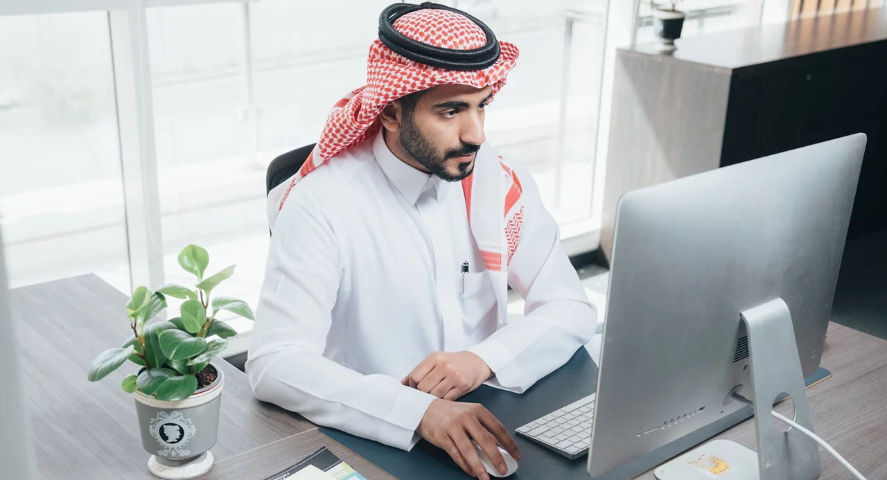 رجل أعمال شرق أوسطي يرتدي زياً تقليدياً يعمل على الكمبيوتر في مكتب حديث مع وجود نبات ووثائق بالقرب منه