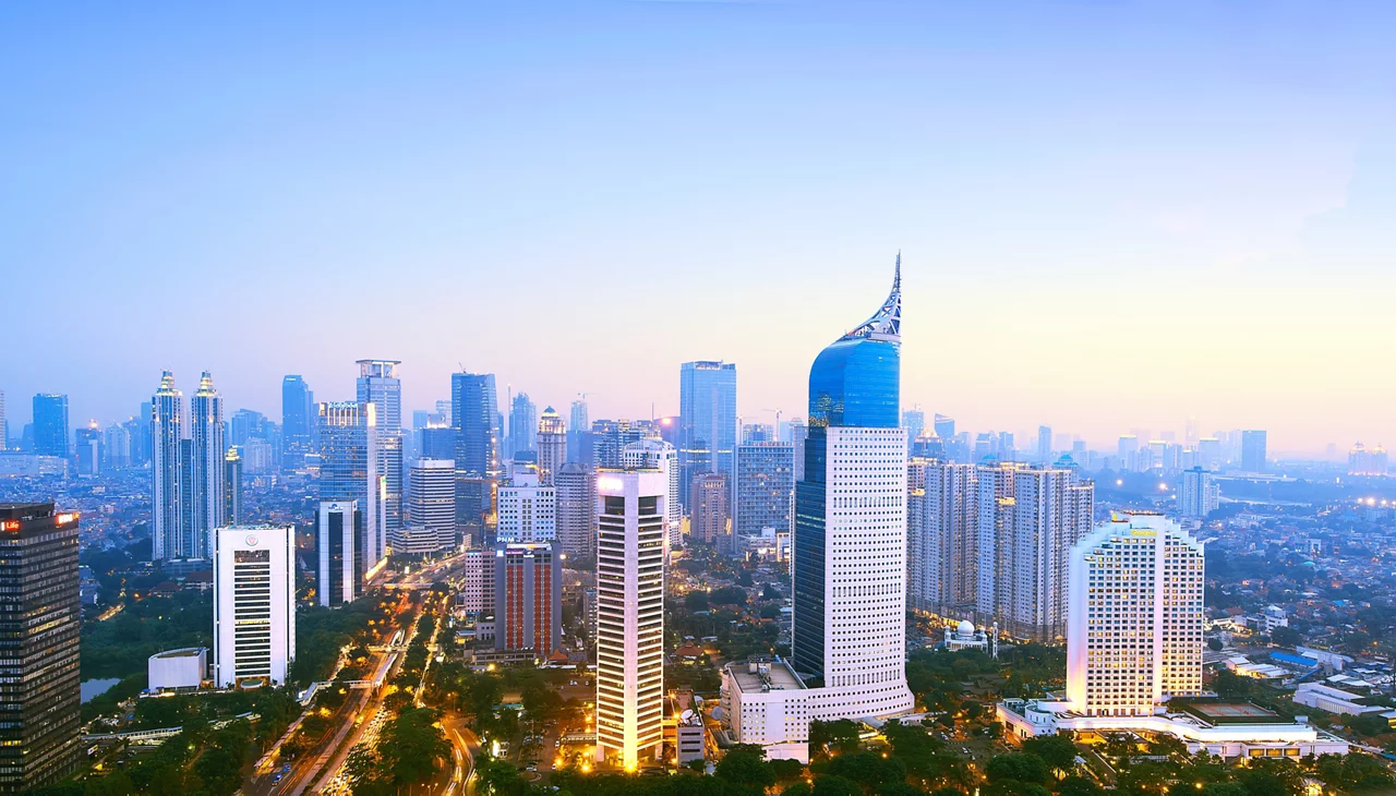 Jakarta skyline at dusk with illuminated skyscrapers including the distinctive blue-topped BNI 46 Tower against a pastel sunset sky.