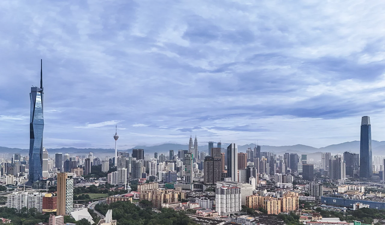 Kuala Lumpur skyline featuring Petronas Towers, KL Tower, and Exchange 106 under cloudy blue skies with mountains in background.