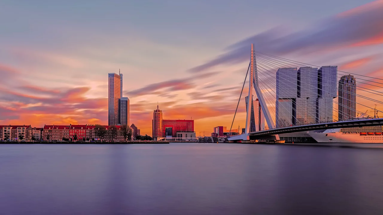 De skyline van Rotterdam bij zonsondergang met de Erasmusbrug en moderne hoogbouw weerspiegeld in kalm water.