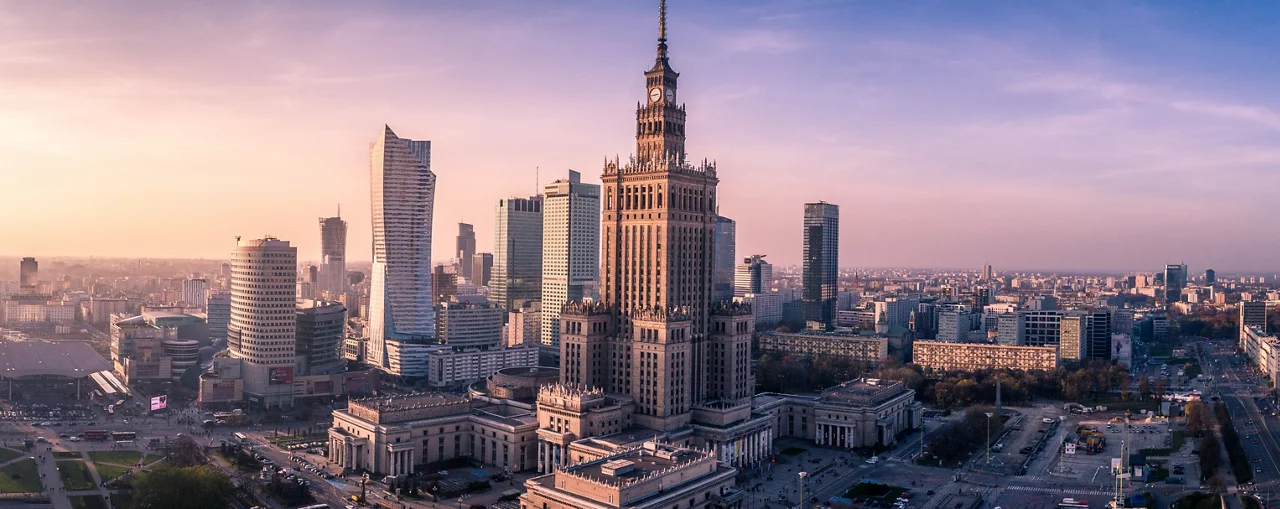 Panoramiczny widok na panoramę Warszawy w Polsce o zachodzie słońca, ukazujący ikoniczny Pałac Kultury i Nauki pośród nowoczesnych wieżowców.