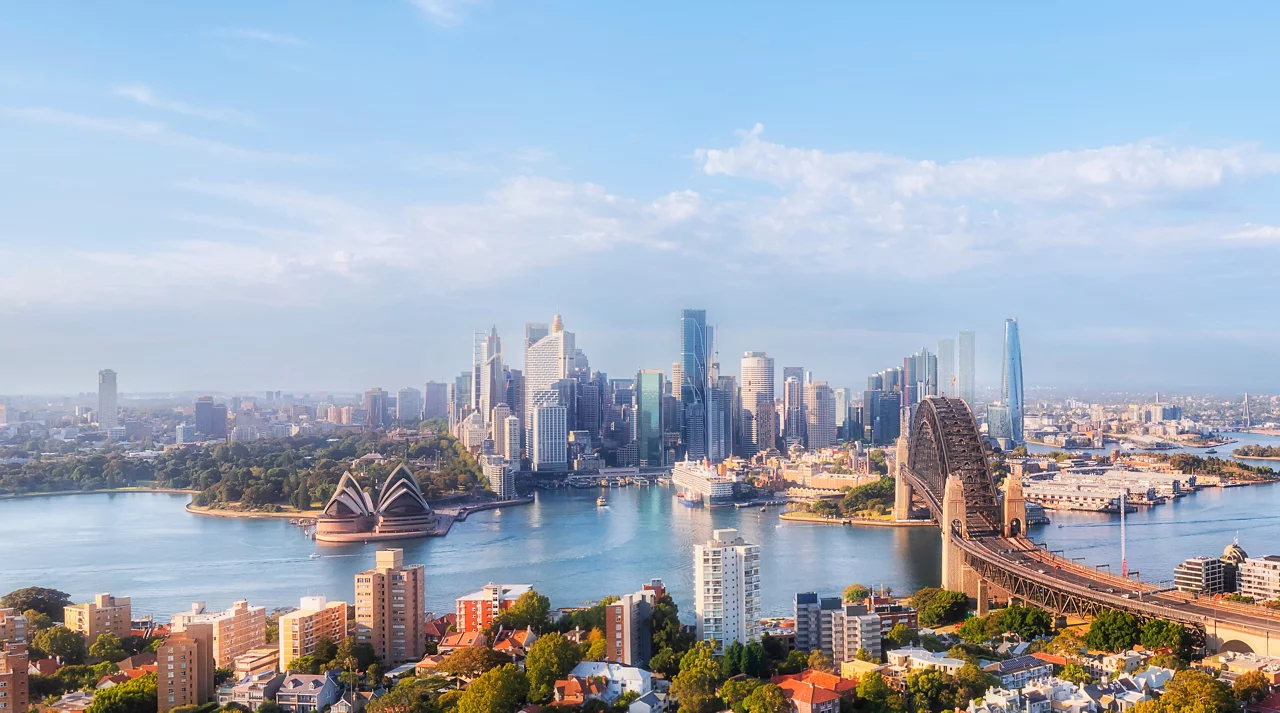 Aerial view of Sydney harbor with Opera House, Harbor Bridge, city skyline, and surrounding residential areas under blue sky.