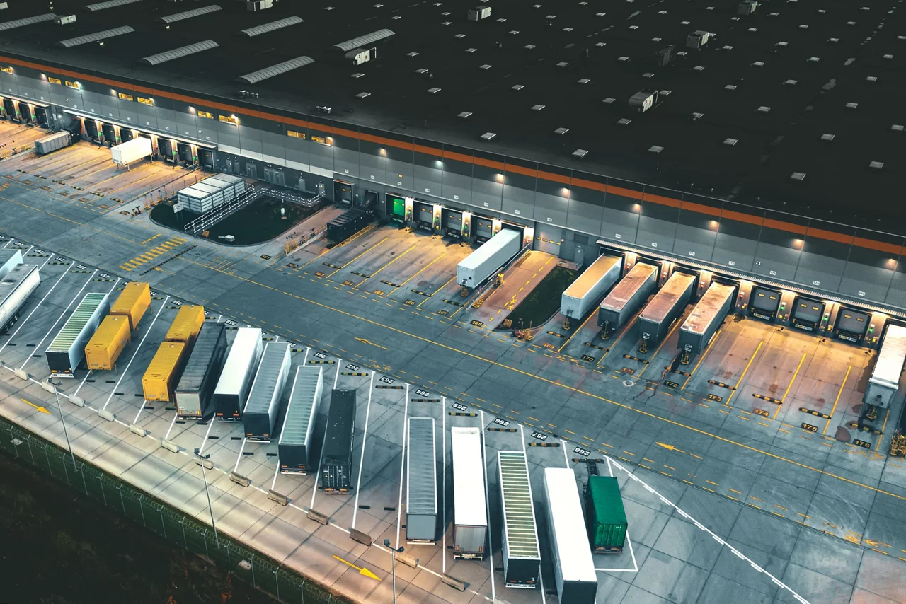 Vista aérea de um grande centro de distribuição com semirreboques acoplados nas plataformas de carga e camiões adicionais estacionados em áreas designadas.