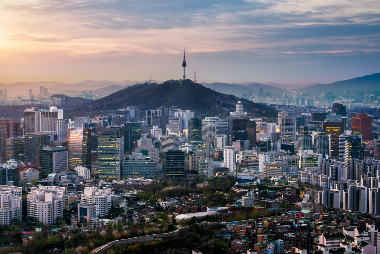 일몰 무렵의 서울 도시 풍경으로, 산 위에 남산타워가 우뚝 서 있고 주변으로 현대적인 고층 빌딩들과 도시 개발이 둘러싸고 있다.