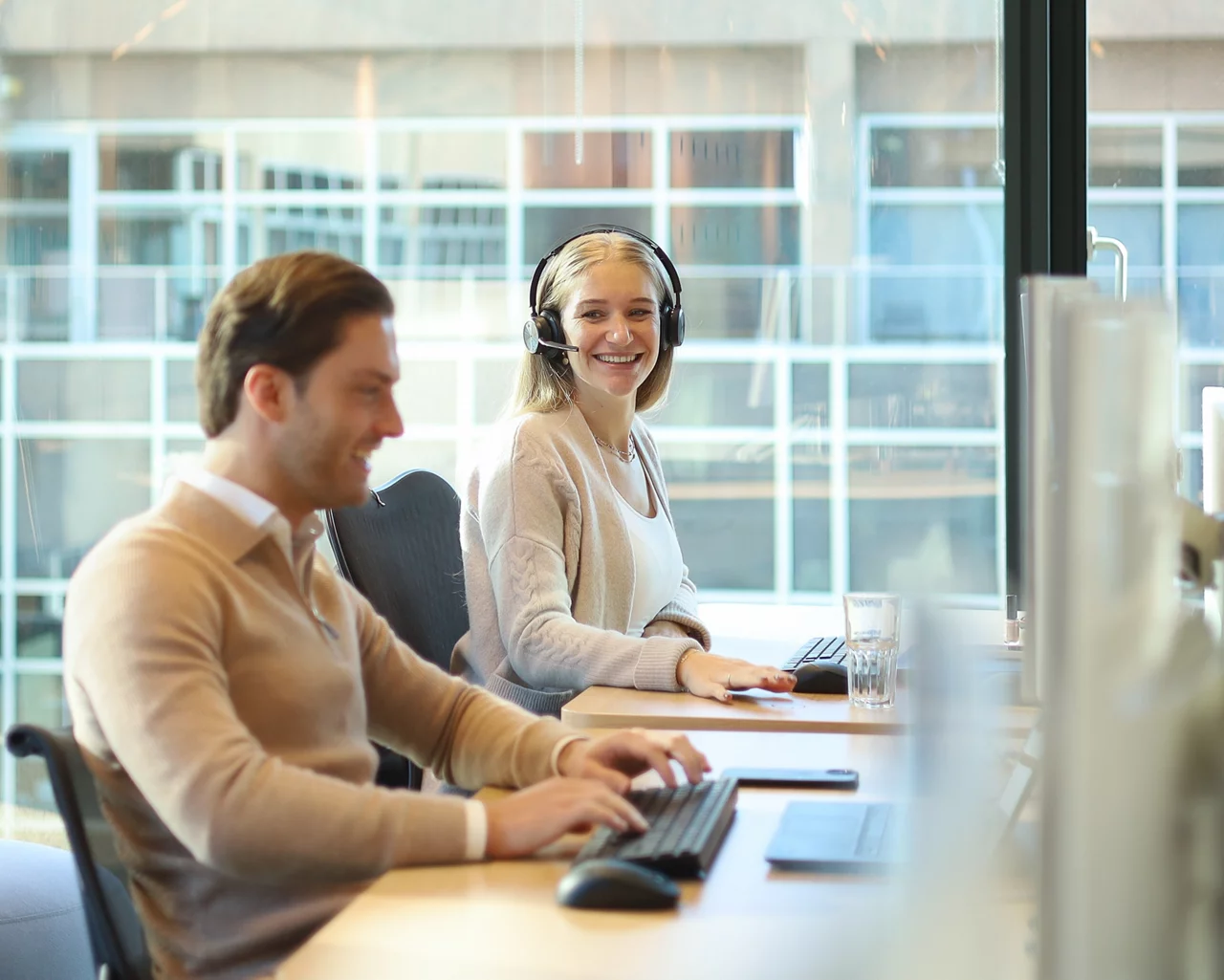 Professional wearing headset smiling while working at desk with colleague in modern office space with glass partitions.