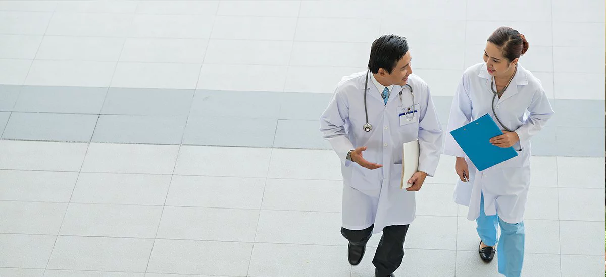 Two doctors in white coats walking and discussing, one holding a blue folder, in a bright tiled hallway or lobby.