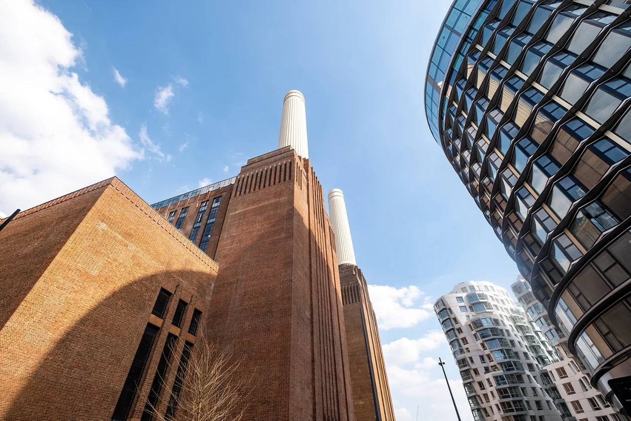 Iconic Battersea Power Station with its chimneys, surrounded by modern glass buildings against a blue sky.