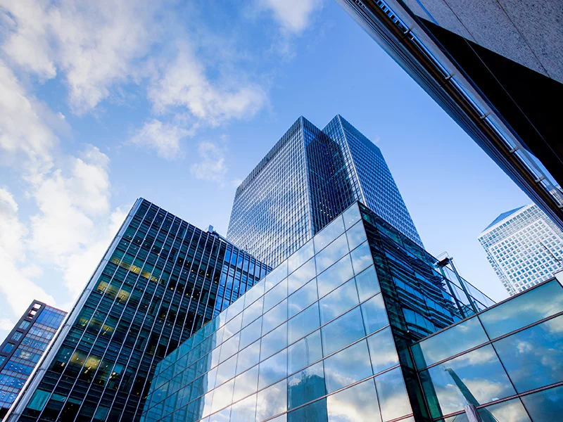 Modern glass skyscrapers with reflective facades against blue sky, viewed from street level in urban financial district.