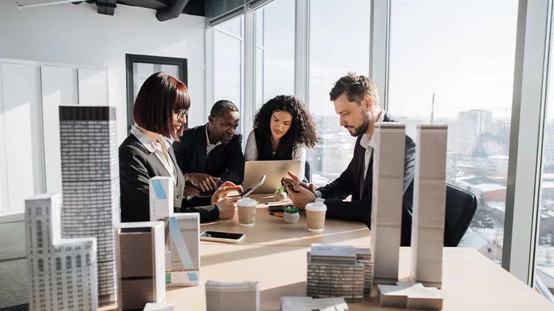Grupo de quatro pessoas trabalha, com celulares e tablet, diante de maquetes de edifícios