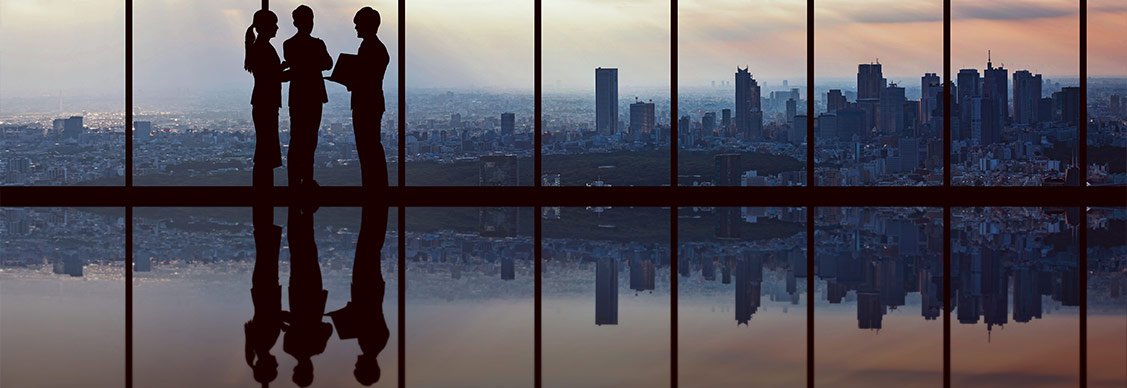 Three business professionals in silhouette stand in conversation near floor-to-ceiling windows