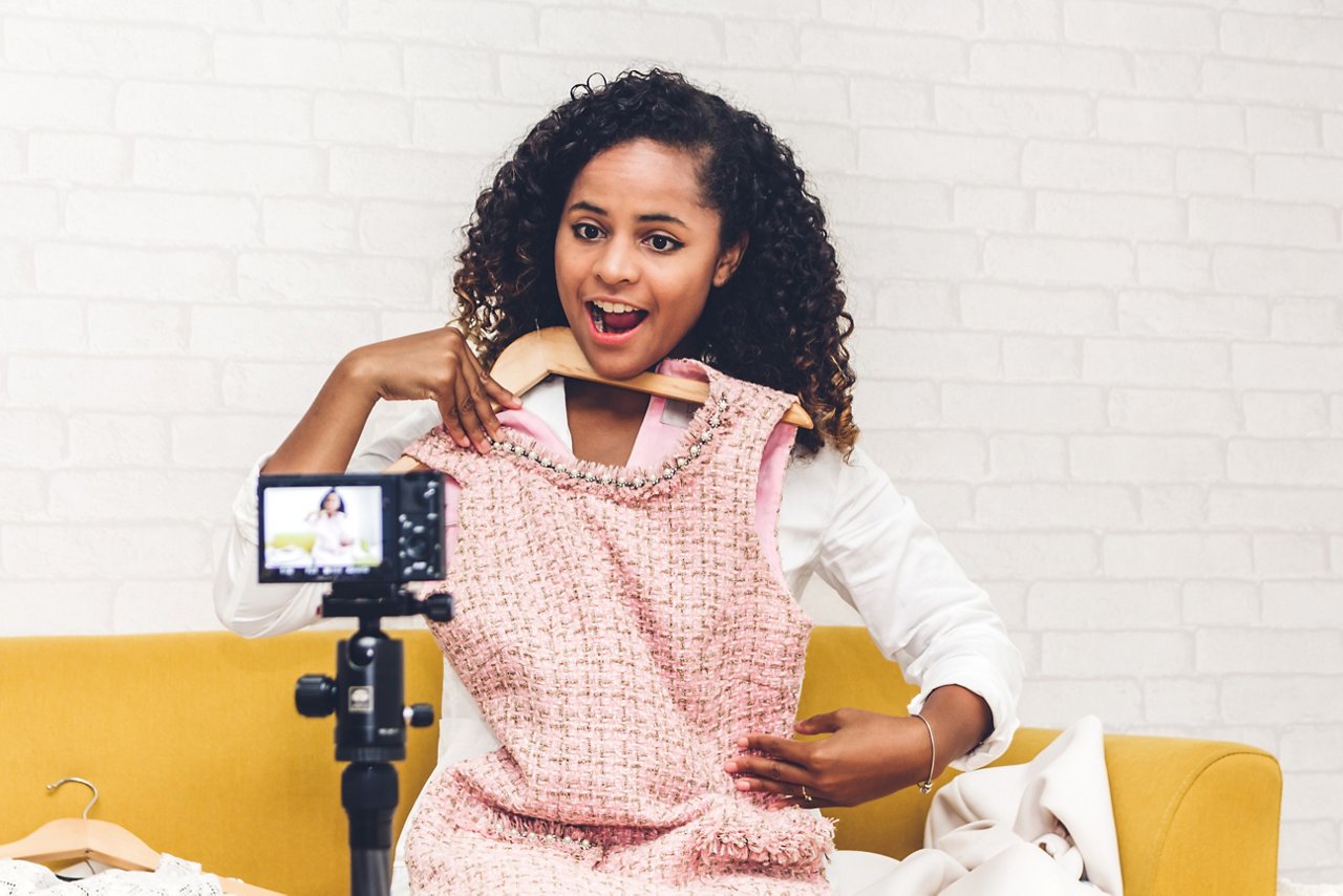 Woman in pink dress recording video on camera while sitting on yellow couch