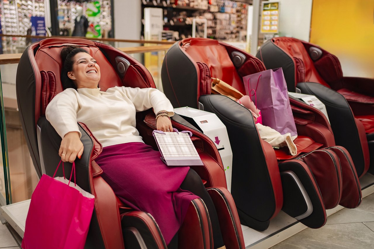 Woman relaxing in red massage chair with shopping bags in electronics store