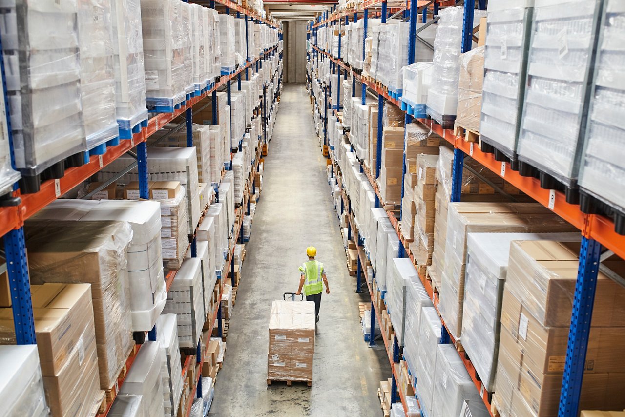 Rear view of male worker pulling cardboard boxes. Employee is walking with pallet jack at aisle. He is working at distribution warehouse.