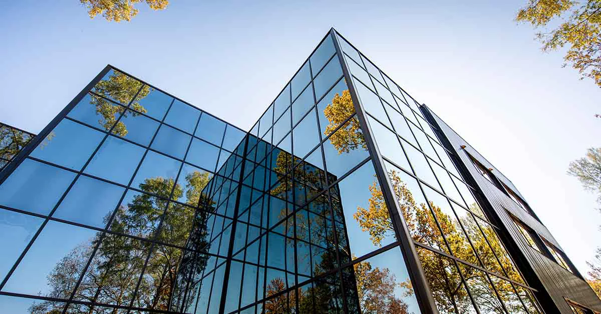 Modern glass office building with a geometric facade reflecting autumn trees against a clear blue sky