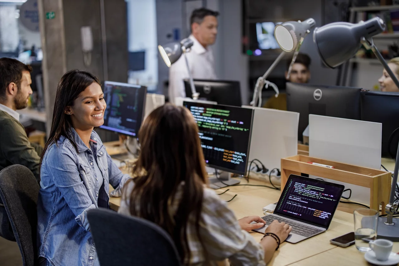 Both women are seated at desks with computers.