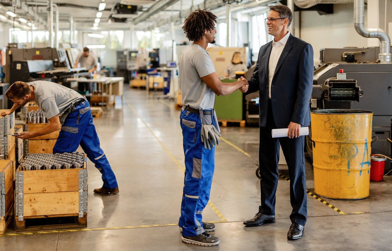 Happy businessman shaking hands with metal worker in industrial facility.