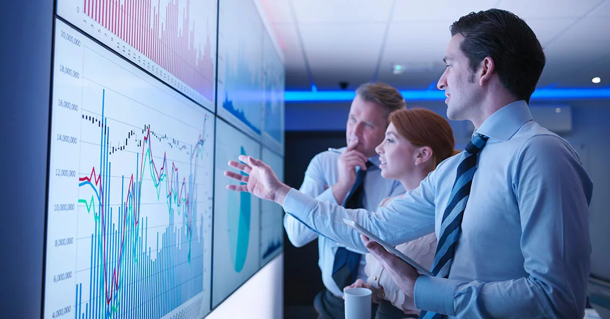 Three business people discuss graphs on screen in meeting room
