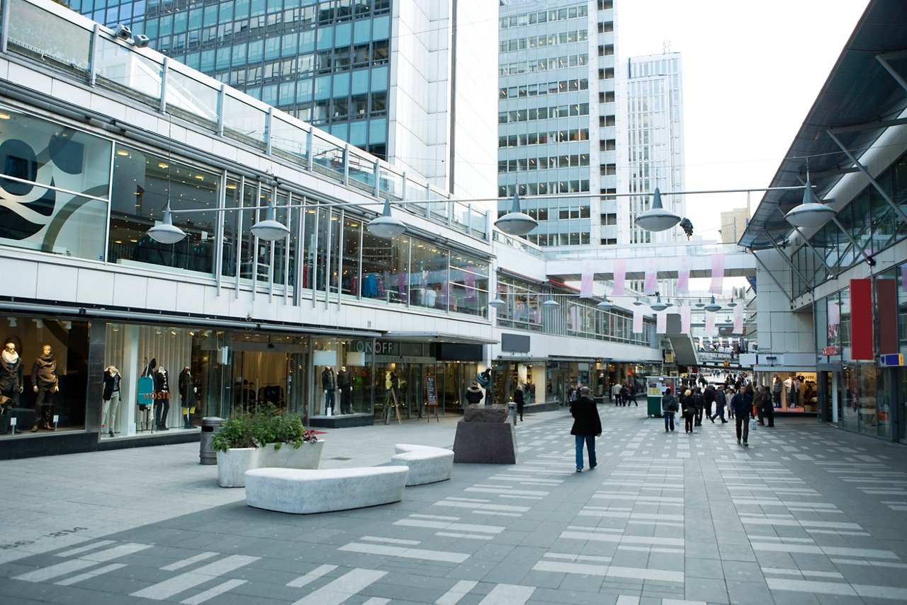 Modern urban shopping plaza with glass storefronts, office towers, and pedestrians walking through paved courtyard.