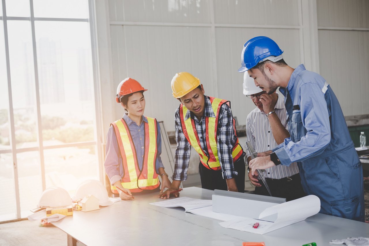 Four construction professionals in hard hats and safety vests collaborate around architectural plans on a table