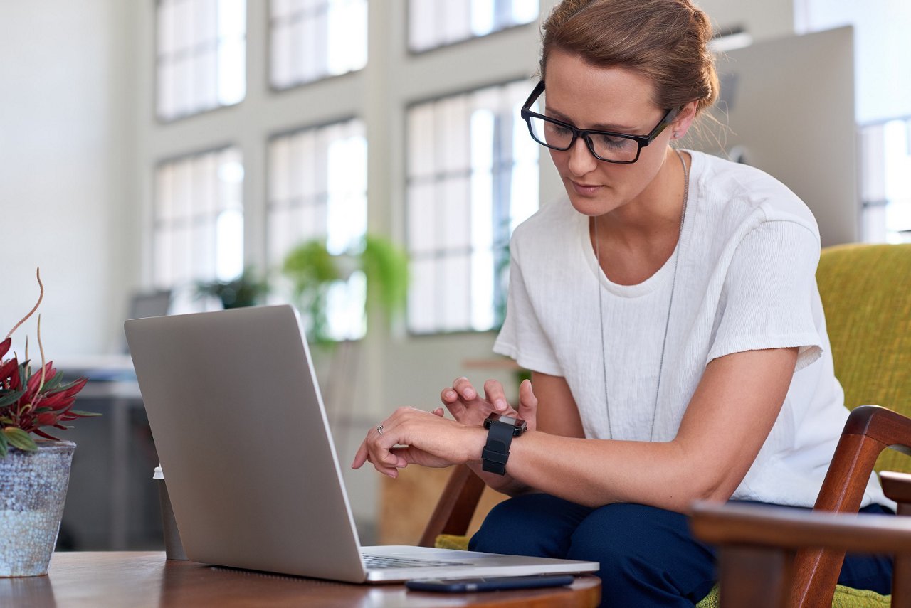 A female sitting on a chair in front of her laptop and accessing her smart watch