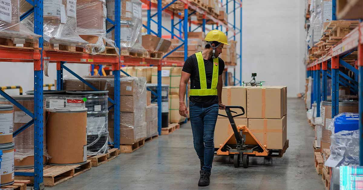 A man wearing a safety vest and hard hat walks through a warehouse.
