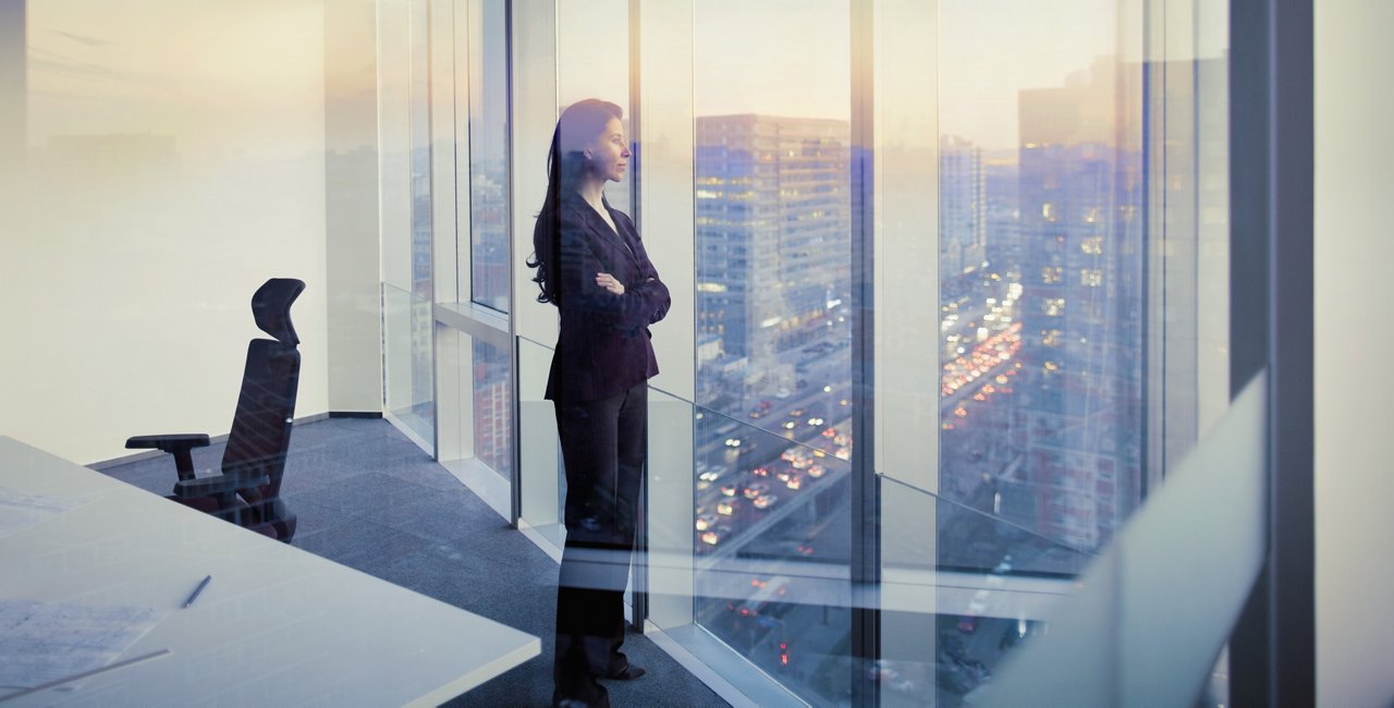 プロフェッショナルなビジネススーツを着た女性が、モダンなオフィスで夕日に照らされた街のスカイラインを見下ろす床から天井までの窓際に立っている。