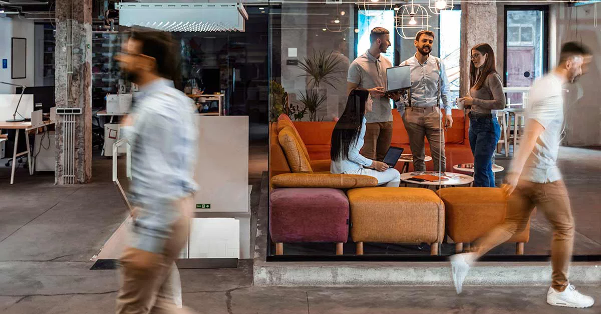 Modern hybrid office with people speaking in a meeting room with a man walking past in front