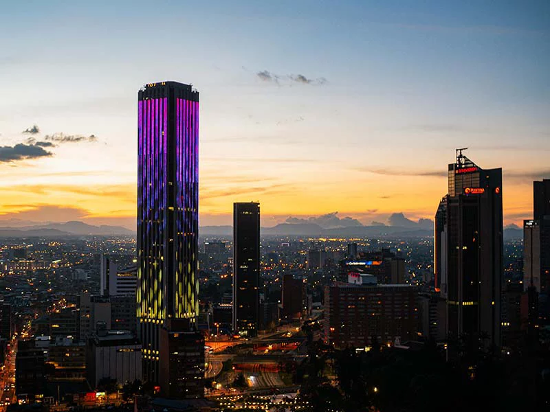 City skyline at sunset with illuminated skyscrapers, featuring a tall purple-lit building against an orange sky.