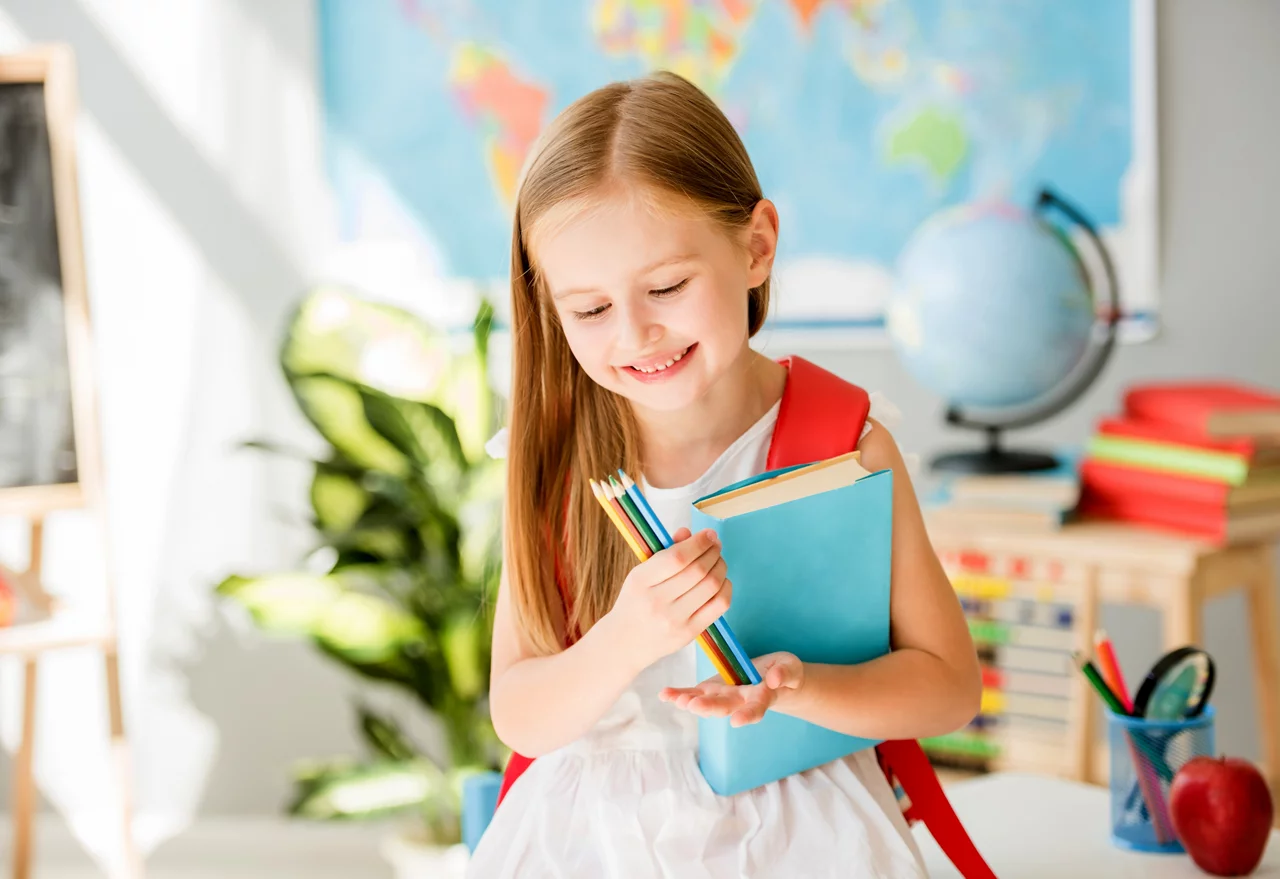 Smiling girl with book and pencils standing in classroom