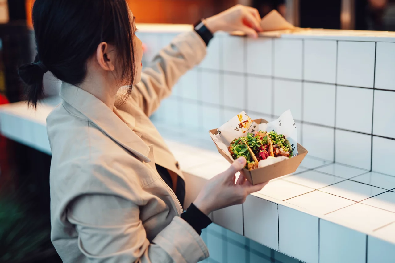 Beautiful young Asian woman holding a box of freshly made Tacos, standing by the food stall. Local foodie experience.