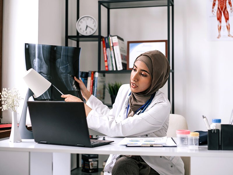 A female doctor wearing a hijab and white medical coat sits at a modern white desk in her office