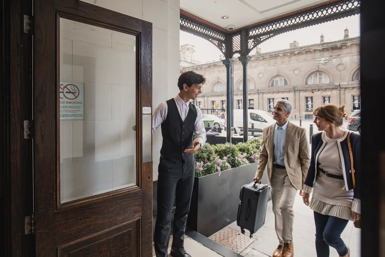 Hotel staff member greets two guests at entrance, historic building facade visible in background.