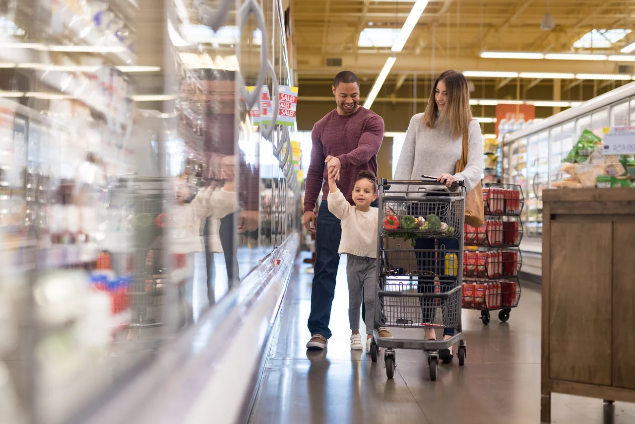A pre-school age girl walks alongside the shopping cart at the grocery store as she helps her parents shop. They're going through the refrigerated section and she is looking up at dad with a mischievous expression.