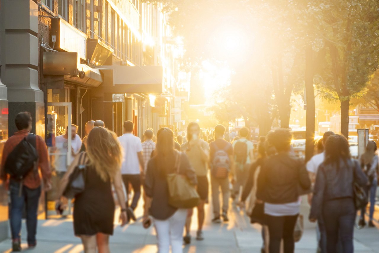 Crowd of anonymous men and women walking down an urban sidewalk with bright glowing sunlight in the background on a busy street in downtown Manhattan, New York City