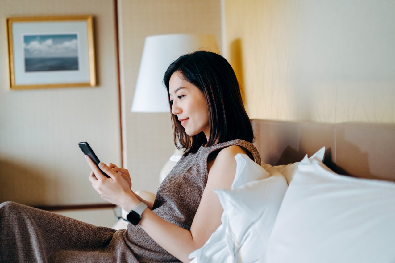 Beautiful young Asian woman using smartphone while lying in bed at cozy home