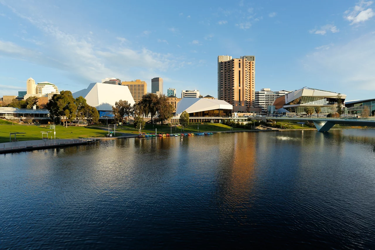 Adelaide cityscape with modern buildings and riverfront, featuring colorful boats on calm water under blue sky.