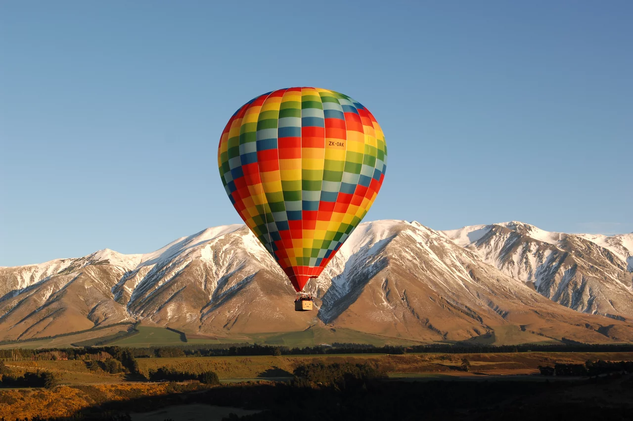 Colorful hot air balloon with rainbow checkered pattern floating over snow-capped mountains and rural landscape.