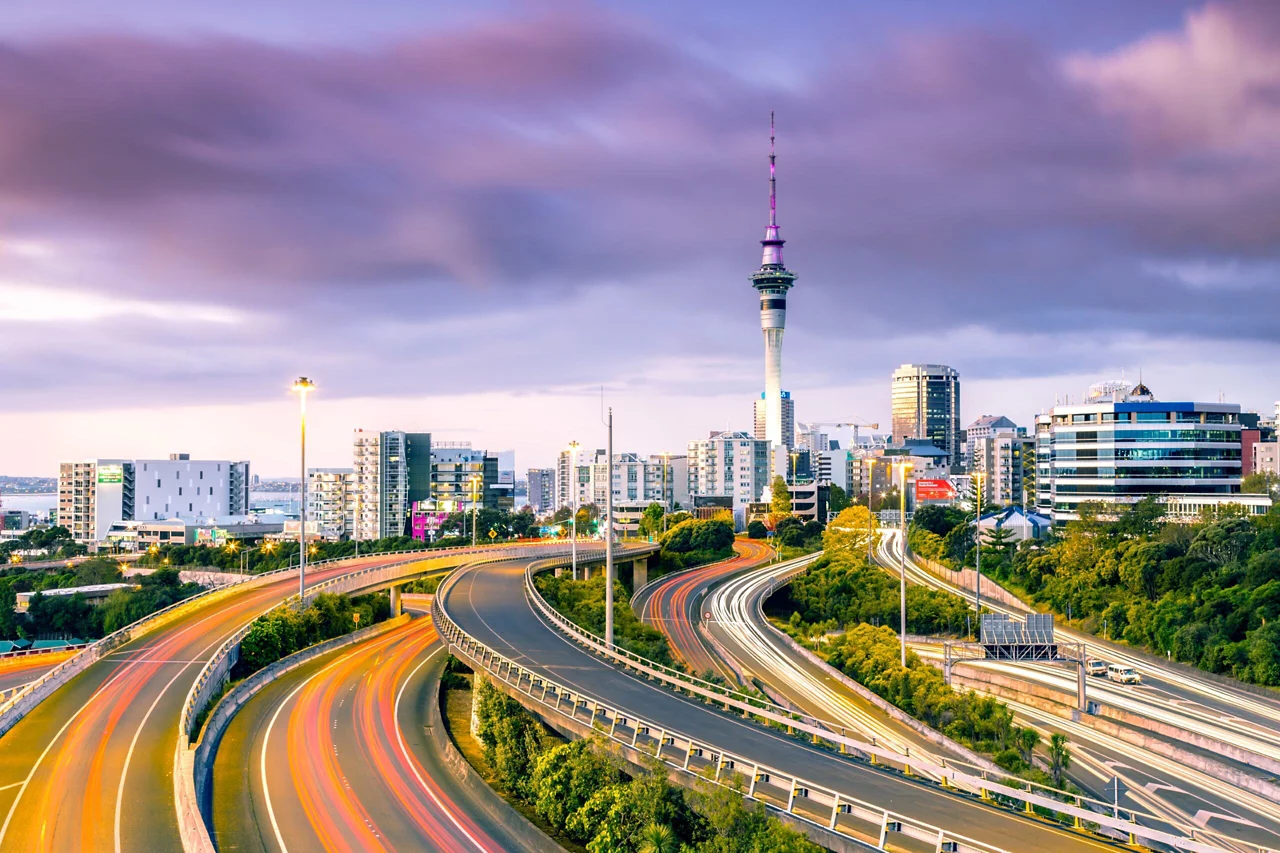 Auckland skyline with Sky Tower and curving highway at dusk, light trails visible on roads leading to city center.
