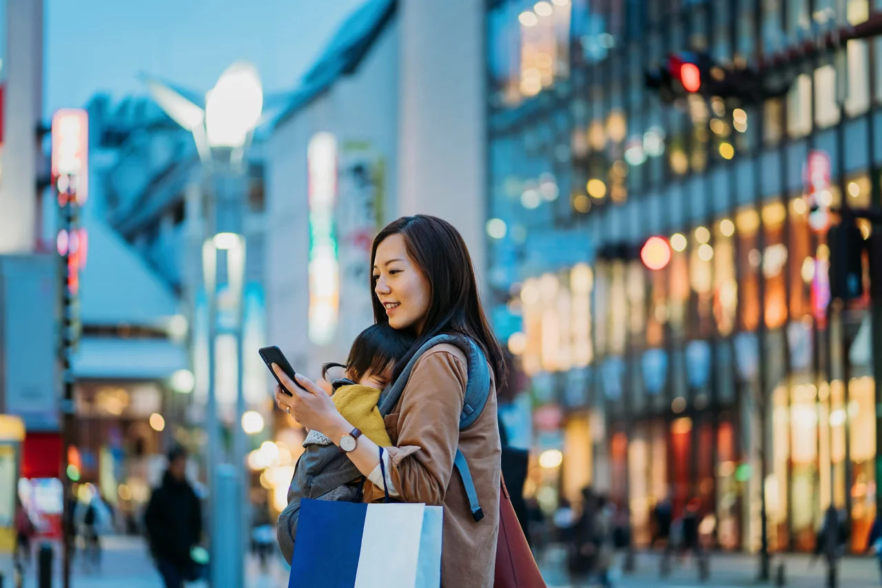 Young Asian mother carrying shopping bag and using smartphone walking in downtown city street shopping with little daughter