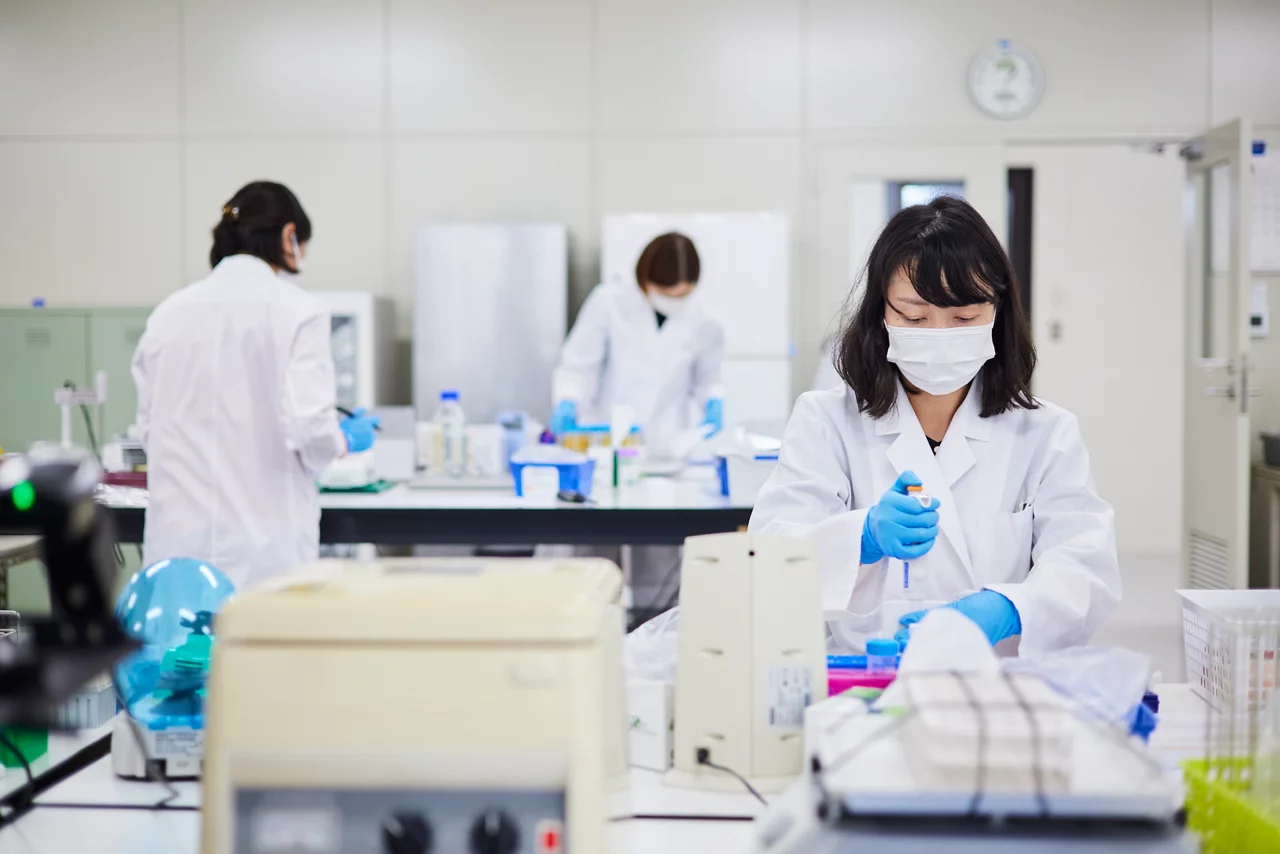 Three lab technicians in white lab coats and masks working in a clinical laboratory with scientific equipment and samples.
