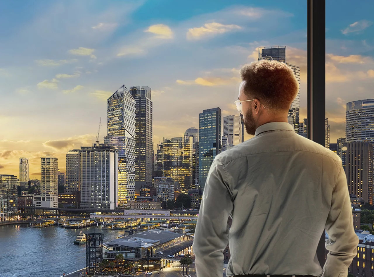 Man overlooking city skyline with tall buildings and waterfront at sunset, illuminated urban landscape view.