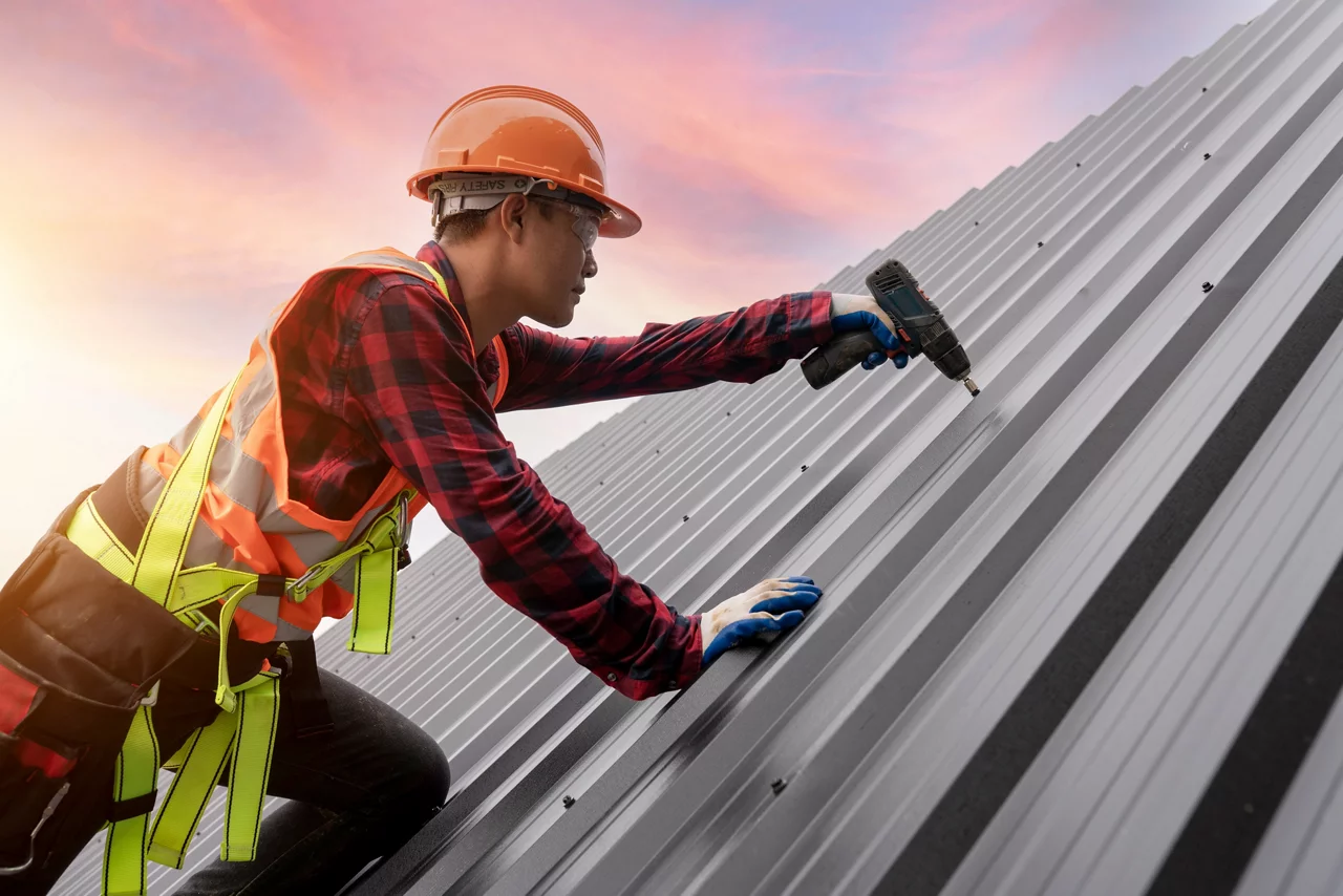 Construction worker in orange hard hat and safety vest installing metal roofing with power drill against pink sunset sky.