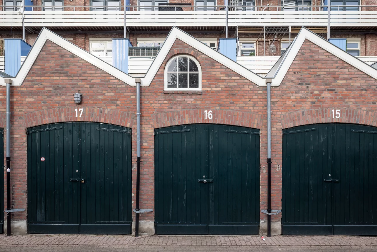 Outdoor street view in front of exterior garage entrance with wooden green doors, brick facade and sawtooth gable roof of European apartment or housing.