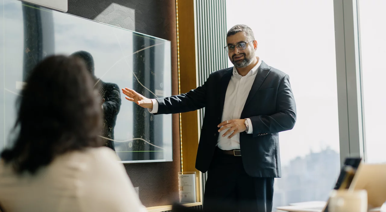 Professional businessman in dark suit presenting to colleagues using wall-mounted display screen in modern office setting.
