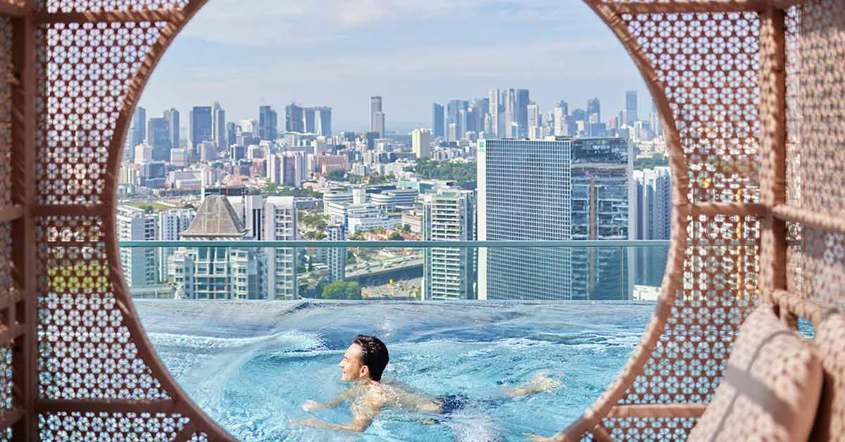A man swims in a rooftop pool, overlooking the city skyline from the top of a building.