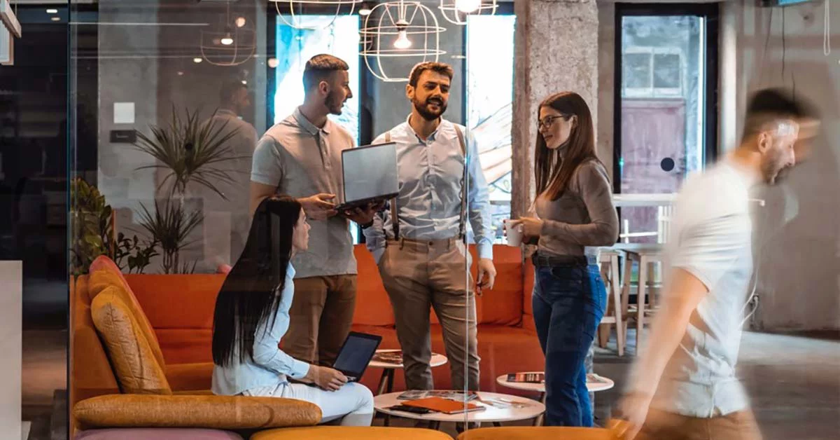 A group of diverse individuals stands in a lobby, engaging with a laptop on a table in front of them.