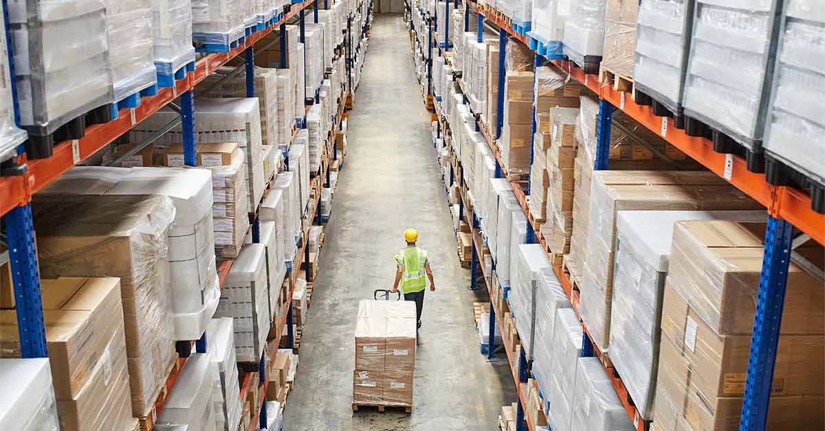 A worker walks through a warehouse aisle, surrounded by shelves filled with various products and supplies.